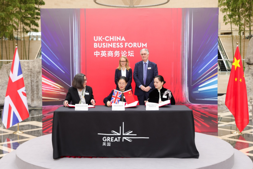 Officials from the UK and China sign agreements at a UK-China Business Forum ceremony with flags flanking the table.