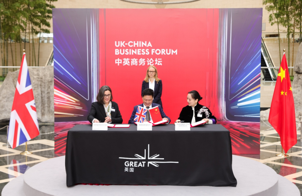 Signatories at a UK–China Business Forum ceremony, three people seated at a black table signing documents, with UK and Chinese flags beside them and a bold red-blue backdrop reading 'UK-CHINA BUSINESS FORUM'.