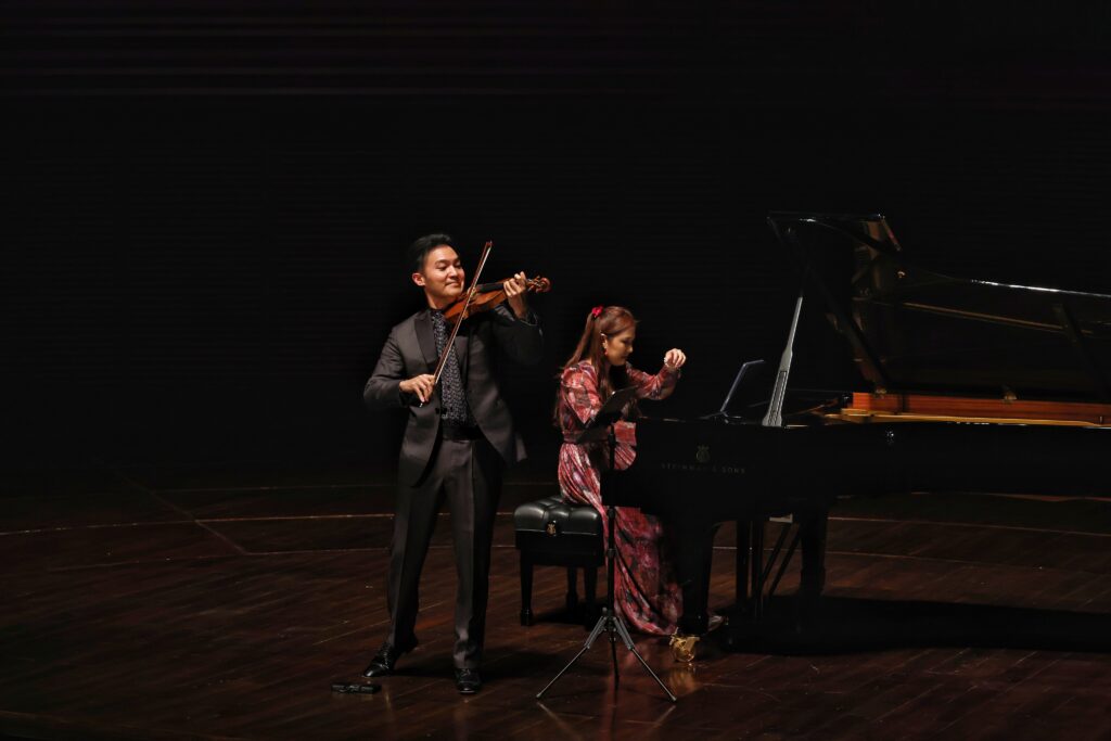 Violinist in a suit plays violin beside a seated pianist on a dark stage with a grand piano nearby