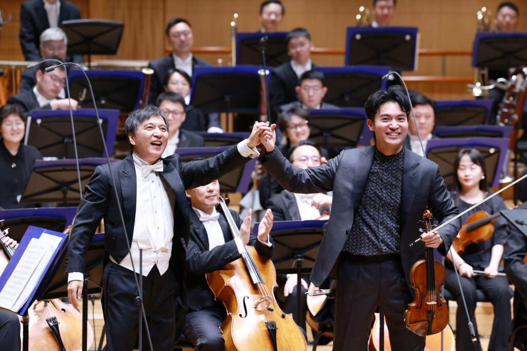 Two conductors in tuxedos raise their hands at the front of a symphony orchestra, with cellos visible in the foreground behind them.