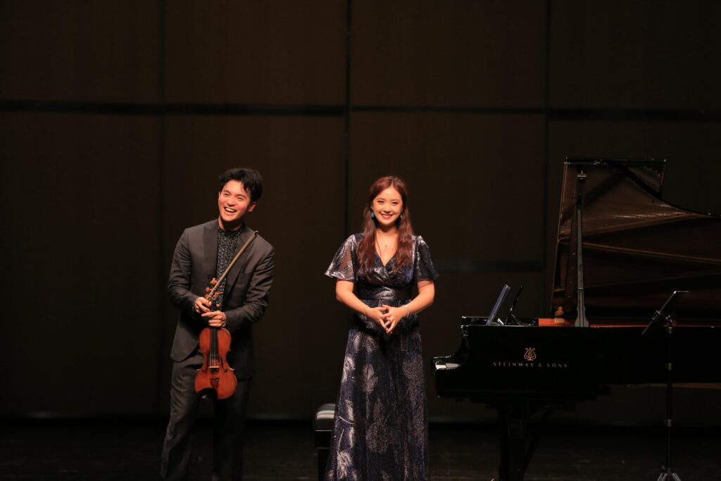 Two musicians on a dim stage: a man with a violin on the left and a woman in a glittery dress beside a grand piano on the right.