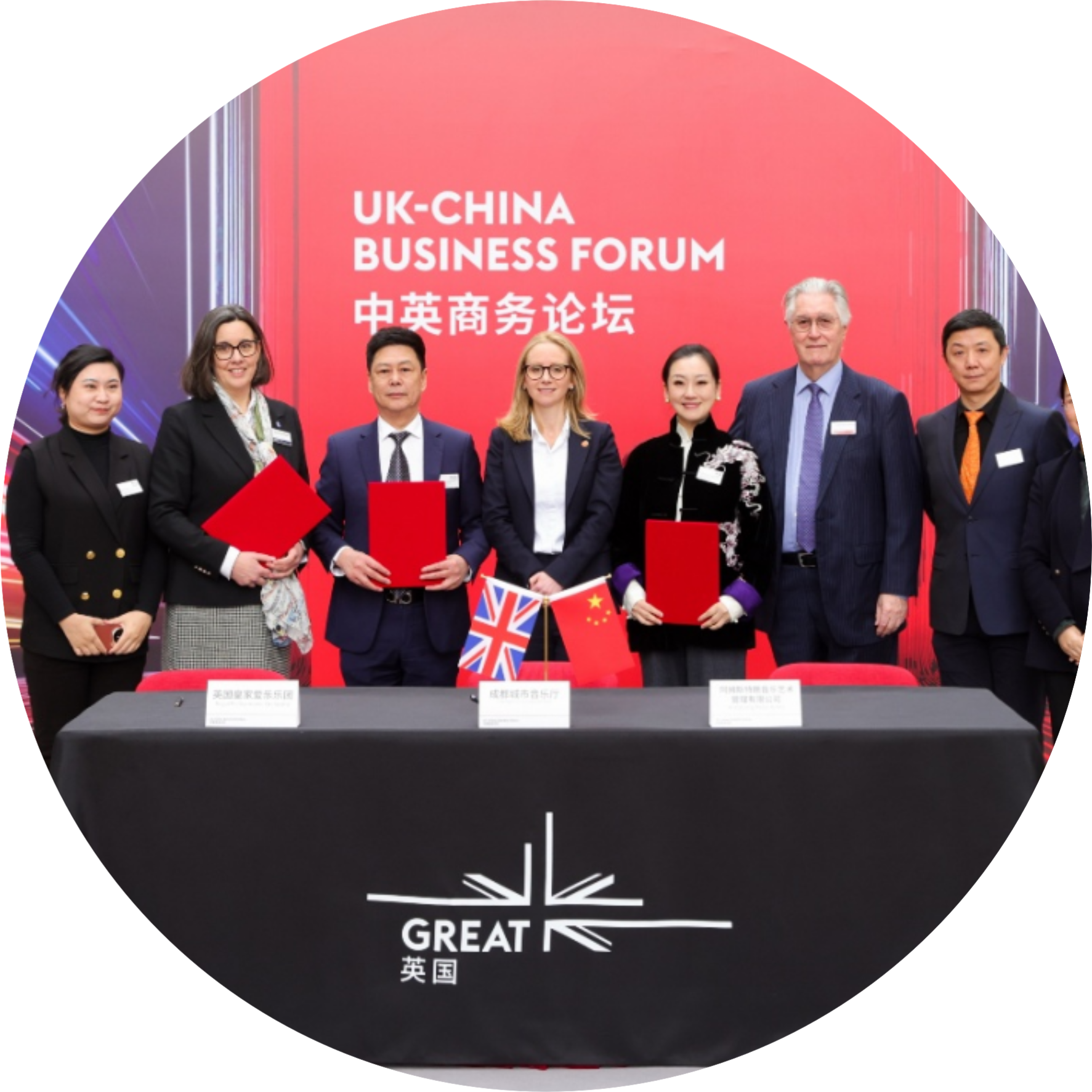 Group of professionals in formal attire posing with red folders at a UK–China Business Forum, red backdrop and small UK/China flags on the table.