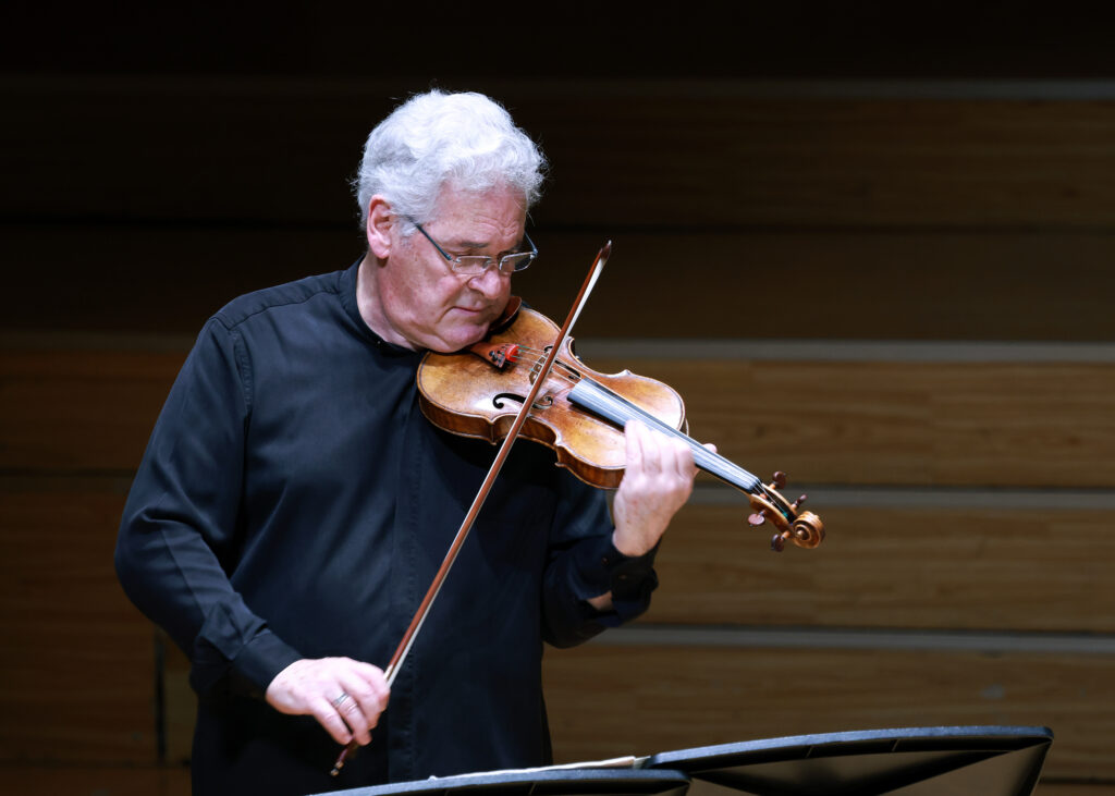 Older man with white hair and glasses playing a violin, wearing a dark shirt, in a wooden-stage setting.