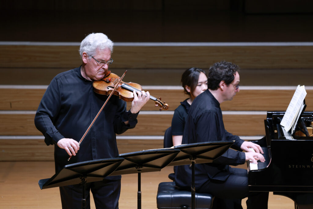 Older man with white hair playing the violin on stage, with a pianist in the background