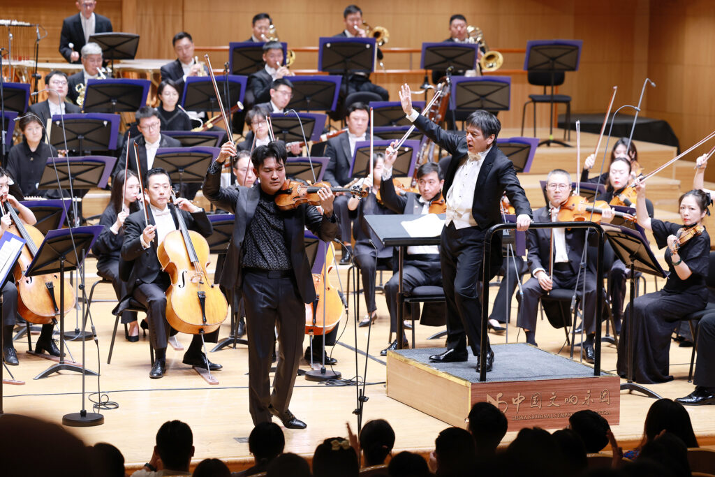 Conductor raises baton as violinists play, front of a large orchestra on a wooden stage.