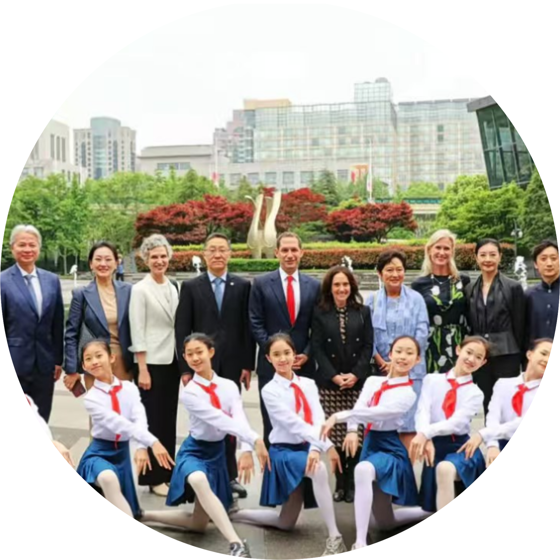 Group of young dancers in white tops and blue skirts performing outdoors, with city buildings and greenery behind them.