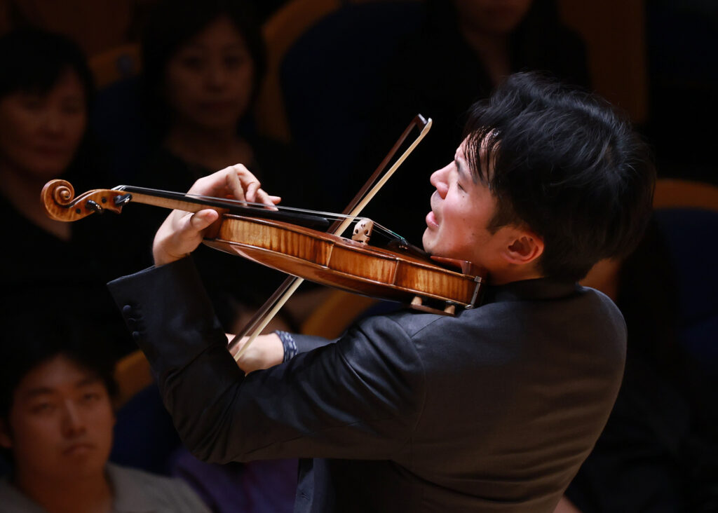 Violinist in a black suit playing a violin, bow drawn across strings with an audience in the dim background.