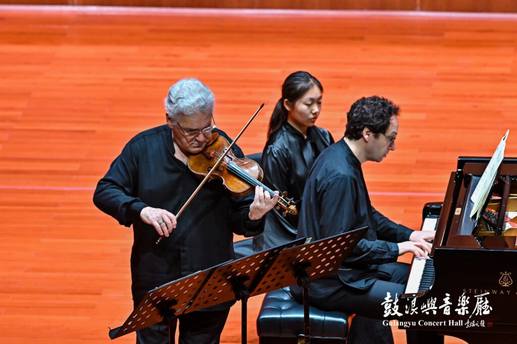 Two musicians perform on a stage: an older man playing violin while a younger pianist accompanies, against an orange wooden backdrop.