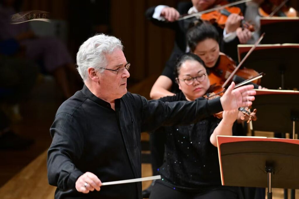 Conductor with white hair in a black shirt directs an orchestra with a baton, while violinists perform behind him.