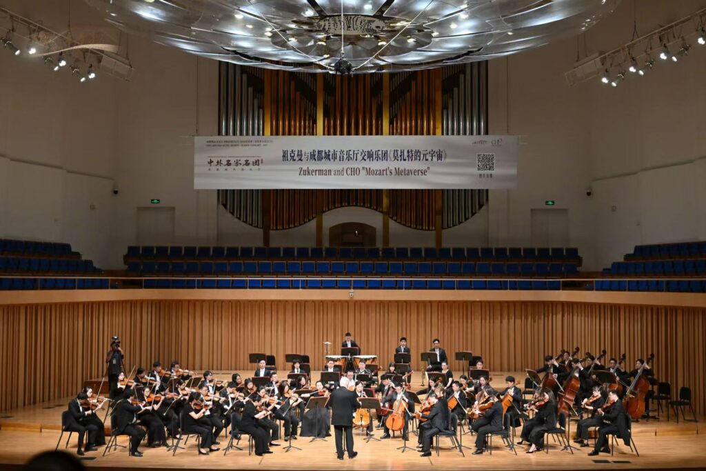 Full orchestra performing on a stage in a concert hall, conductor at front with musicians playing string and wind instruments; a large banner hangs above the stage.