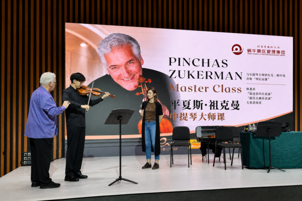 Violinist and student on stage with a mentor watching, a large banner reads 'Pinchas Zukerman Master Class' in the background.
