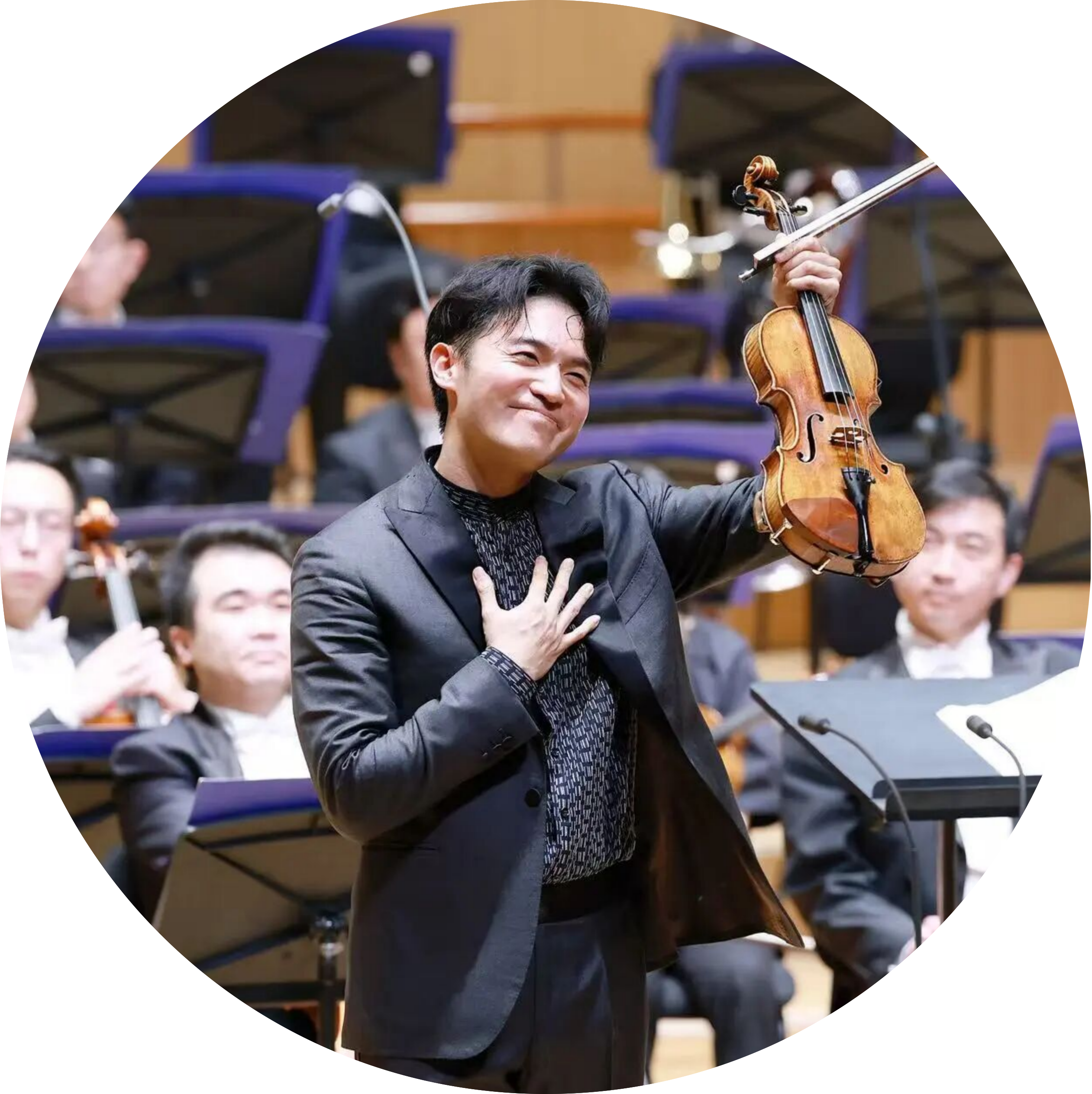 Smiling violinist in a black suit performs in an orchestra in a concert hall behind him.